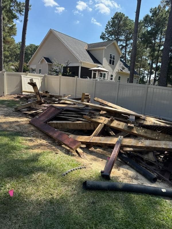 A pile of discarded wooden deck debris sits in a grassy backyard in front of a white vinyl fence and a two-story house.