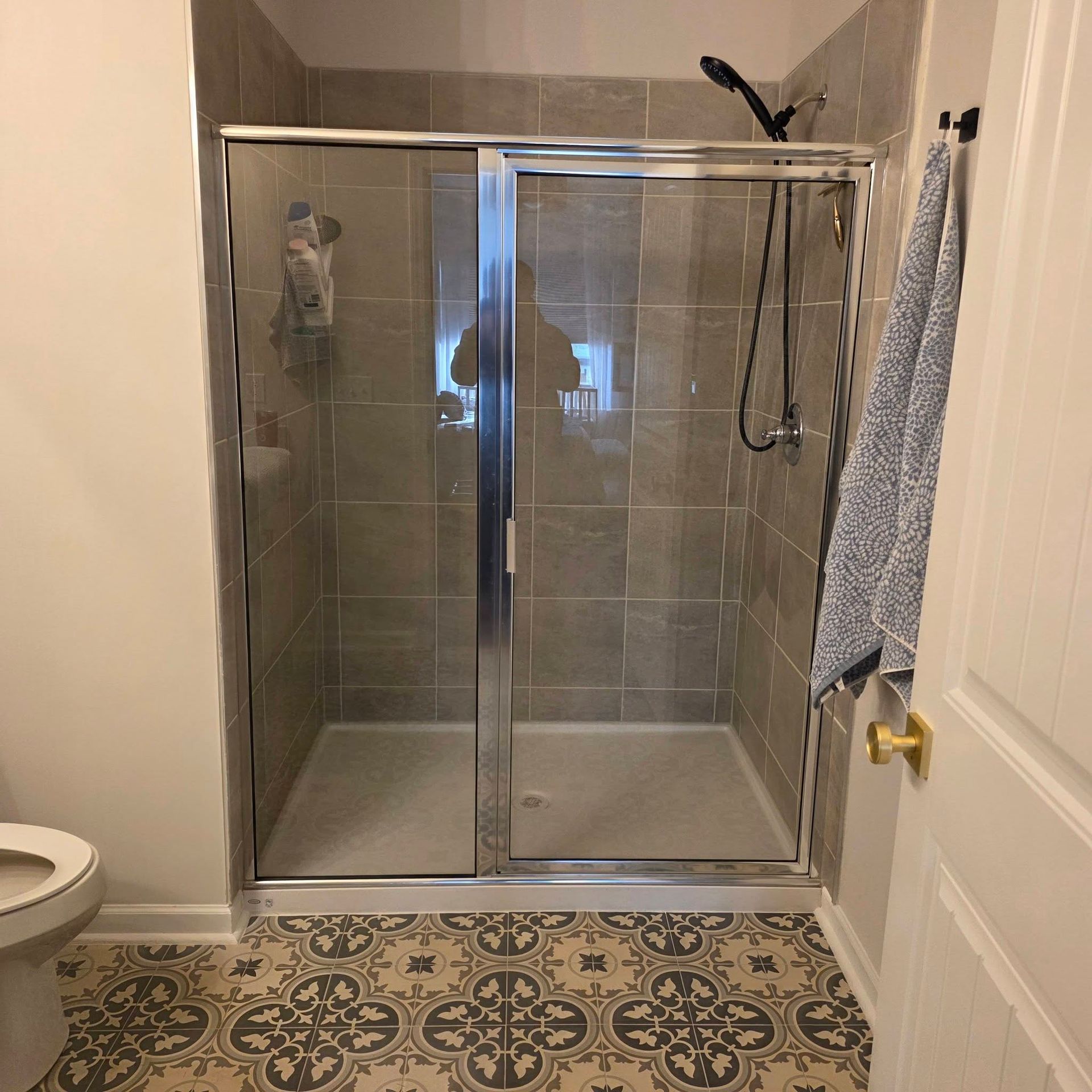 A bathroom with a tiled walk-in shower, glass doors, and patterned tile flooring. A towel hangs on a door handle nearby.