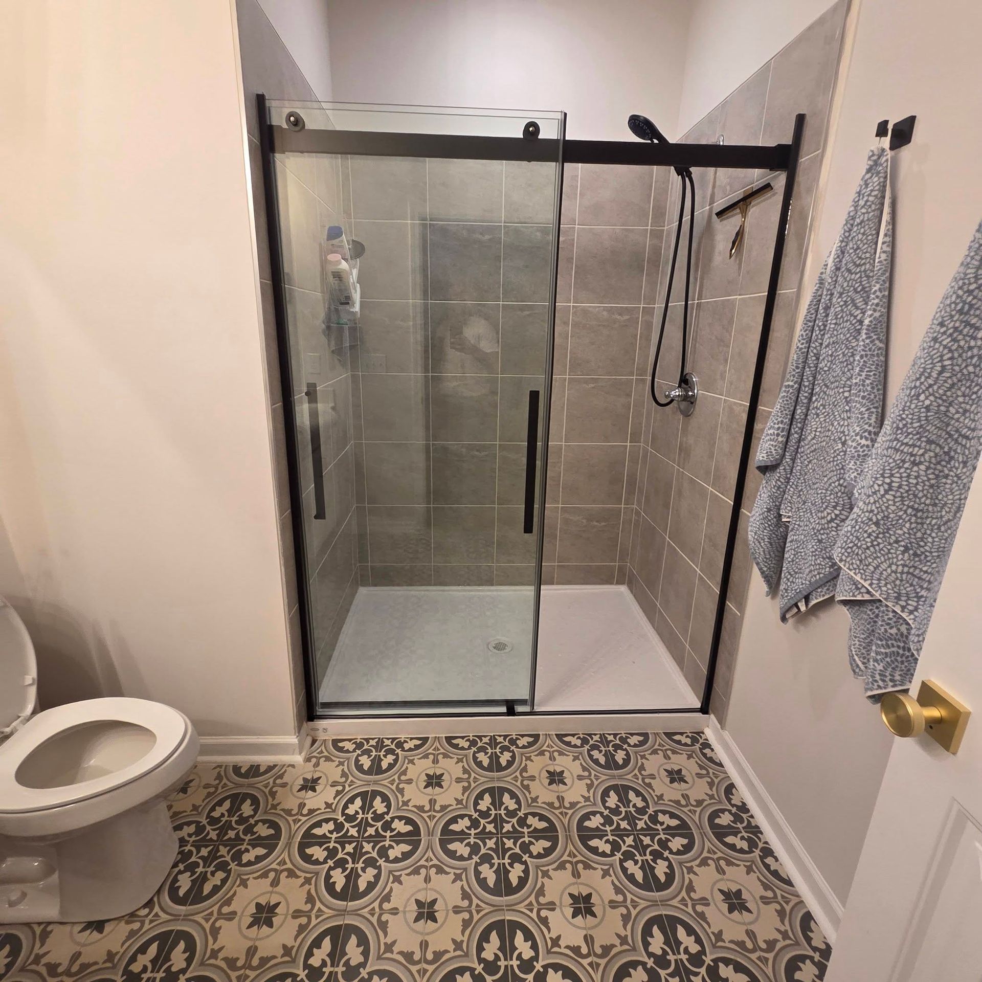 A bathroom with a sliding glass door shower, gray subway tiled walls, patterned tile floors, and a toilet to the left.