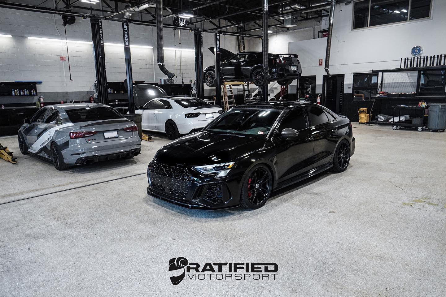 Black sports car in garage, mechanic working on it, another car on a lift. | Ratified Motorsport