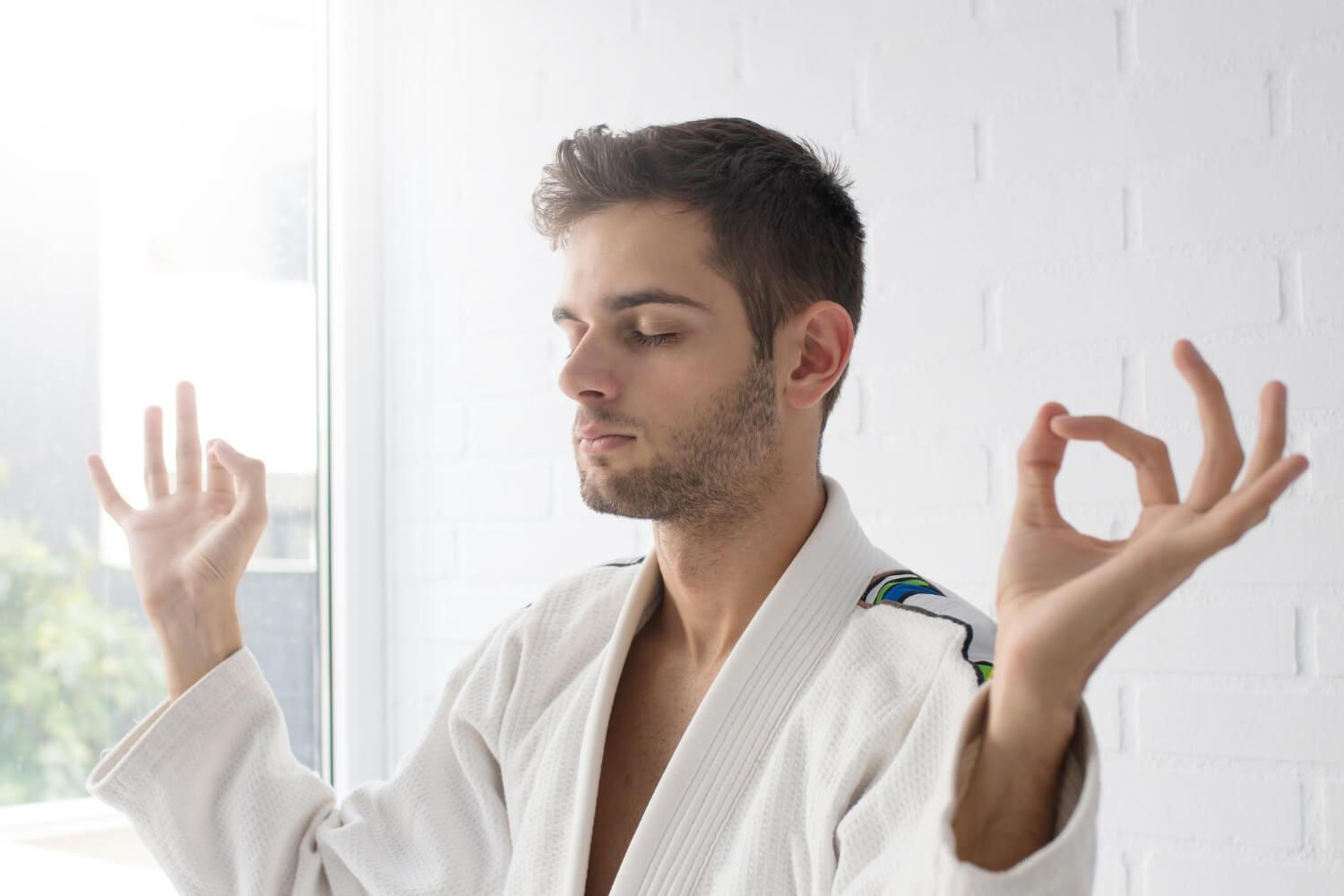 Young man in white robe sitting in meditation pose with hands forming mudra gestures, eyes closed peacefully in bright room with natural lighting