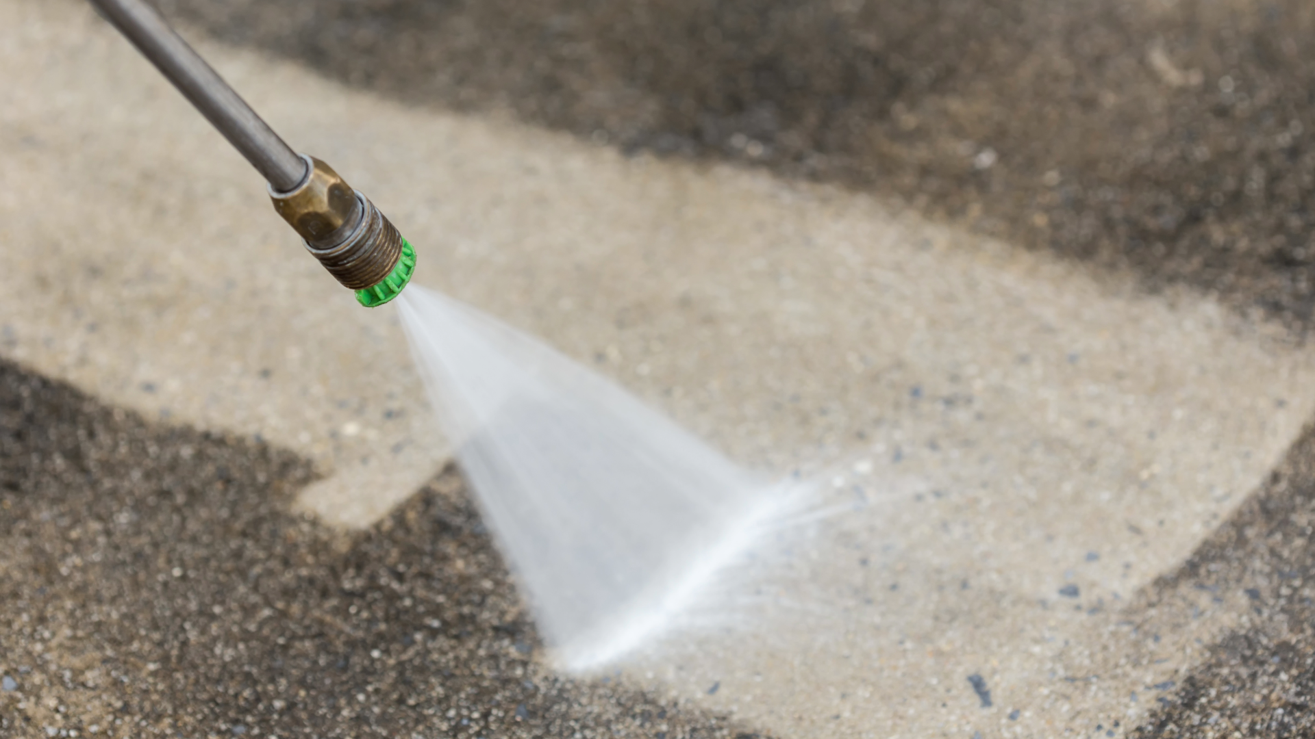 A pressure washer nozzle sprays a fan of water onto a concrete surface, cleaning a dark, stained area to reveal light gray.