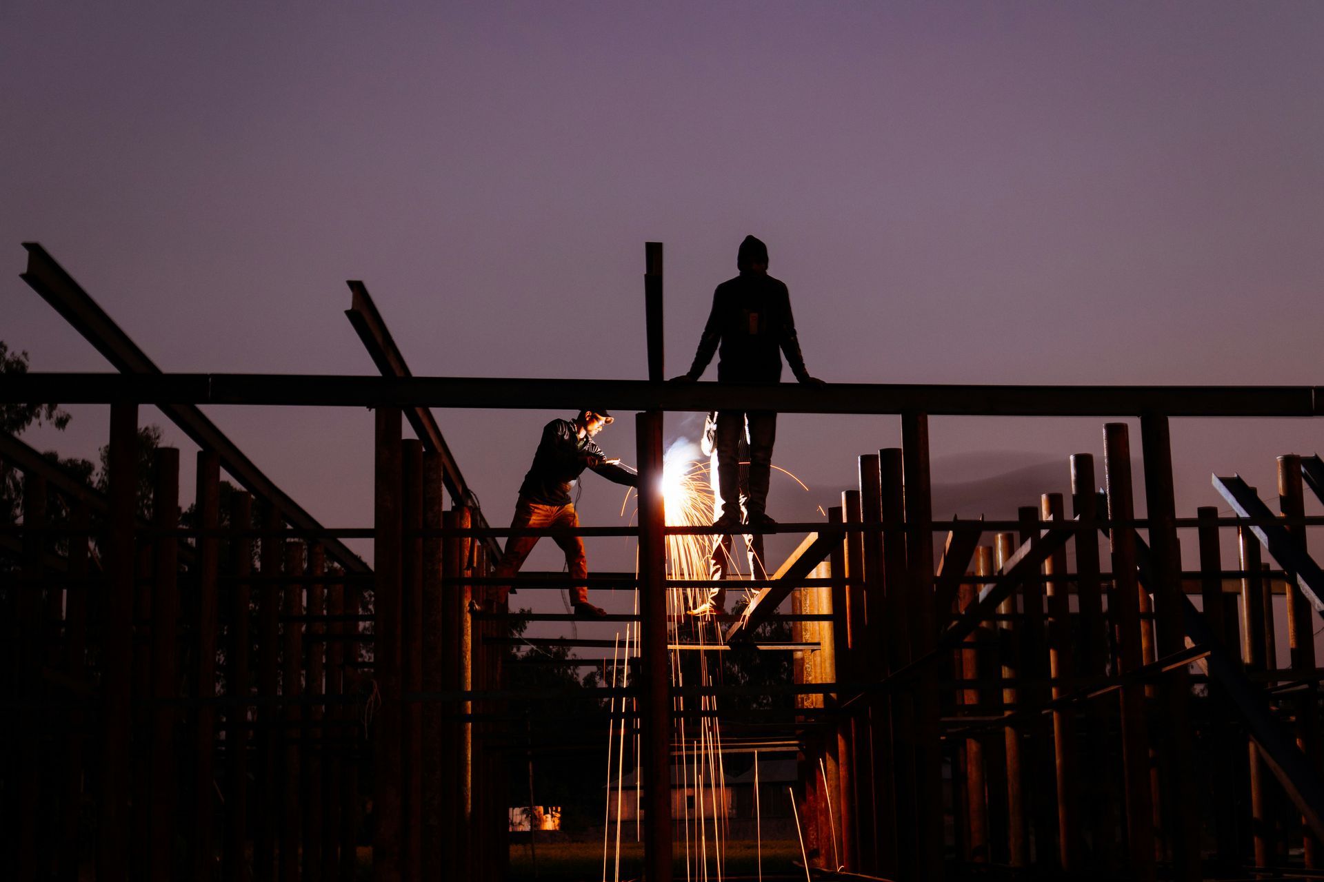 Silhouetted construction workers welding metal beams at dusk. One stands, another welds with sparks.