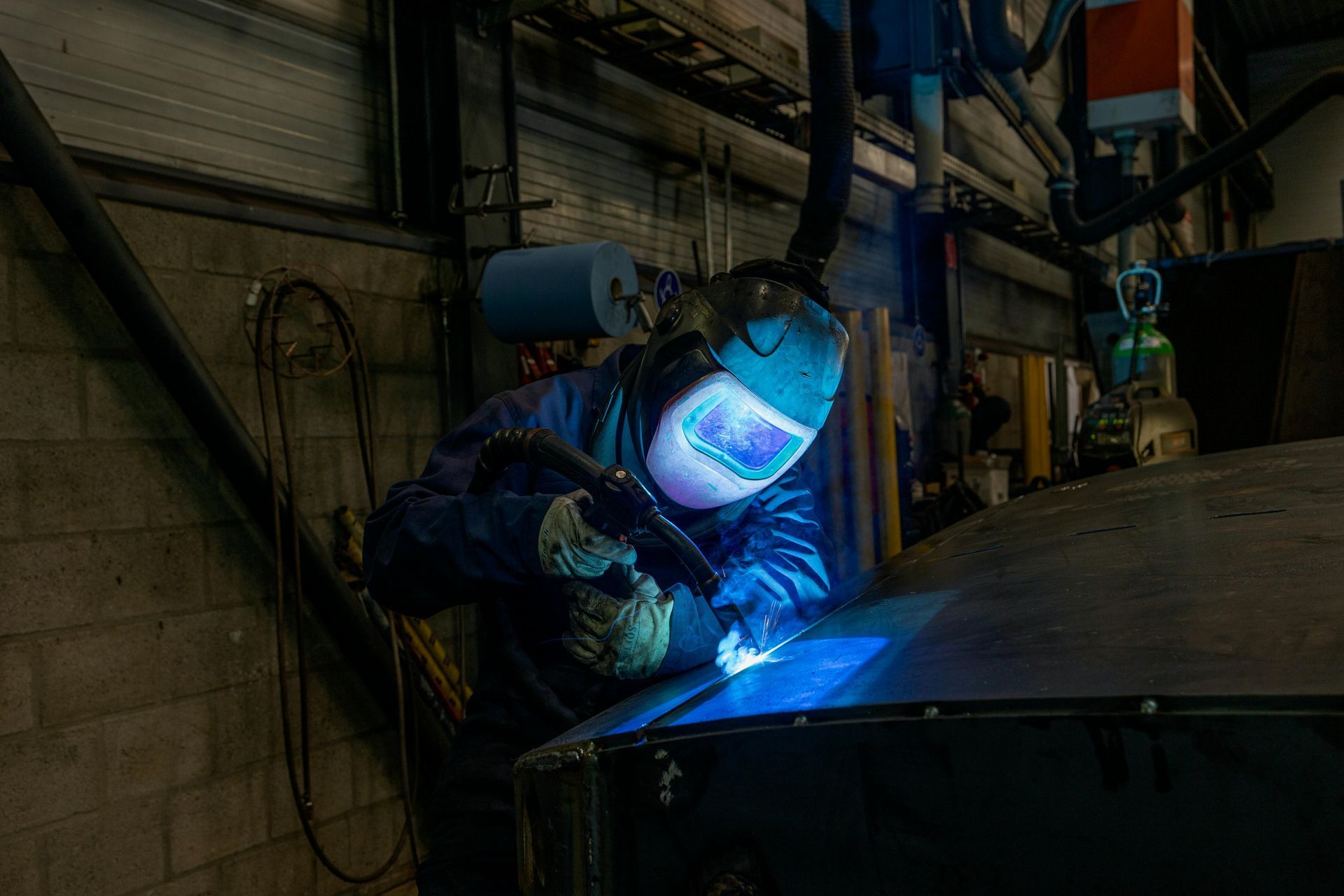Welder wearing a protective mask welding metal in a workshop, bright blue light visible.