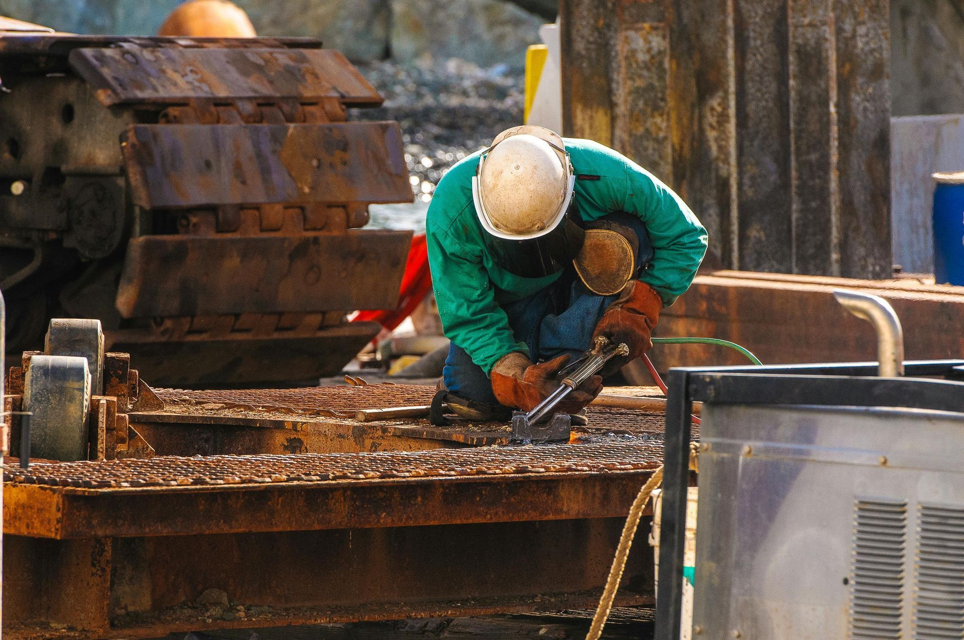 A construction worker in green jacket and hard hat cuts metal with a torch.
