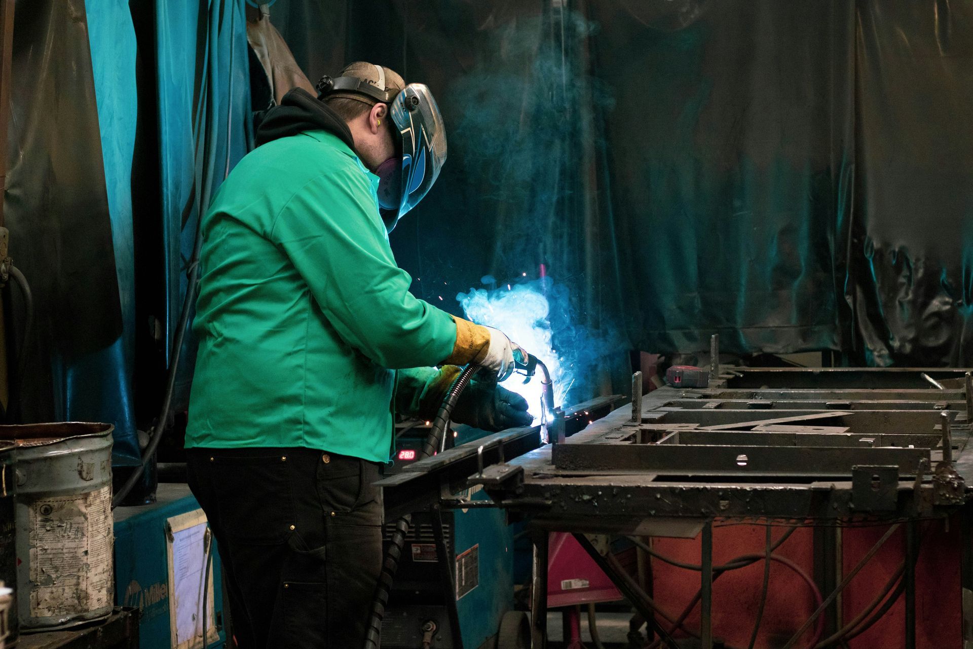 A person in a welding mask, wearing a green shirt, welding metal in a workshop.
