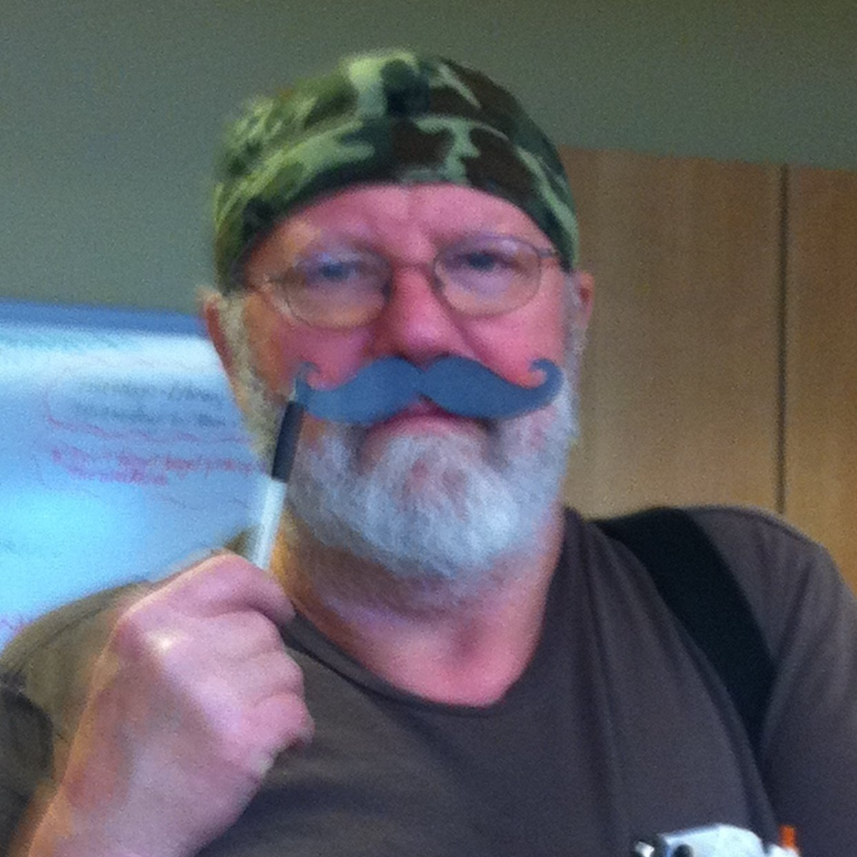 A headshot of Bob Bradbury, a white man with a grey beard and wire rimmed glasses. He is wearing a camo beanie and holding up a fake black mustache.