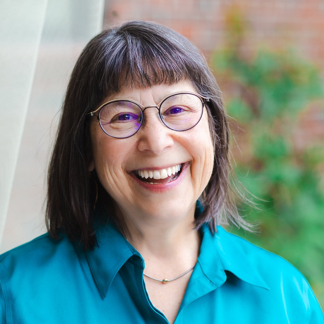 A headshot of Rabbi Braun, a white woman with shoulder length dark hair and bangs wearing a teal collared shirt and round glasses