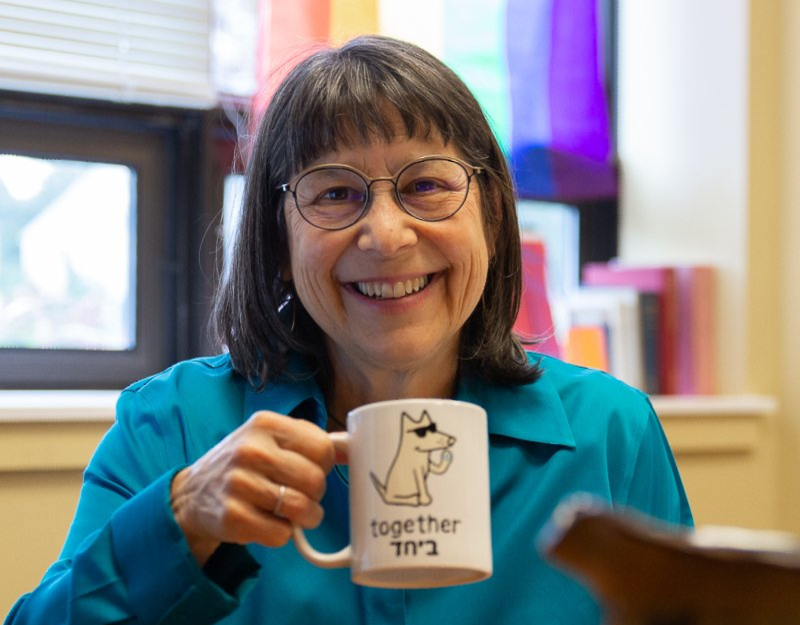 A headshot of Rabbi Braun, a white woman with shoulder length dark hair and bangs wearing a teal collared shirt and round glasses. She is holding a white mug that has a cartoon dog on it with 