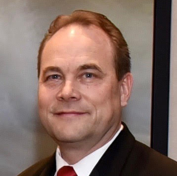 Headshot of Gregory Smestad, a white man with short brown hair. He is wearing a dark suit jacket, a white button-up, and a red tie.