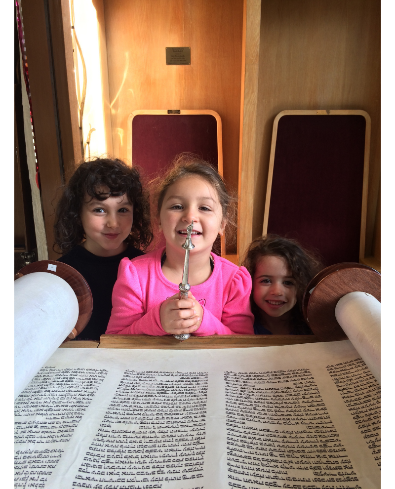A photo of three, white lower elementary aged kids standing in front of an open Torah. The kid in the middle is holding a yad, pointed upwards. They all have long brown hair and are smiling