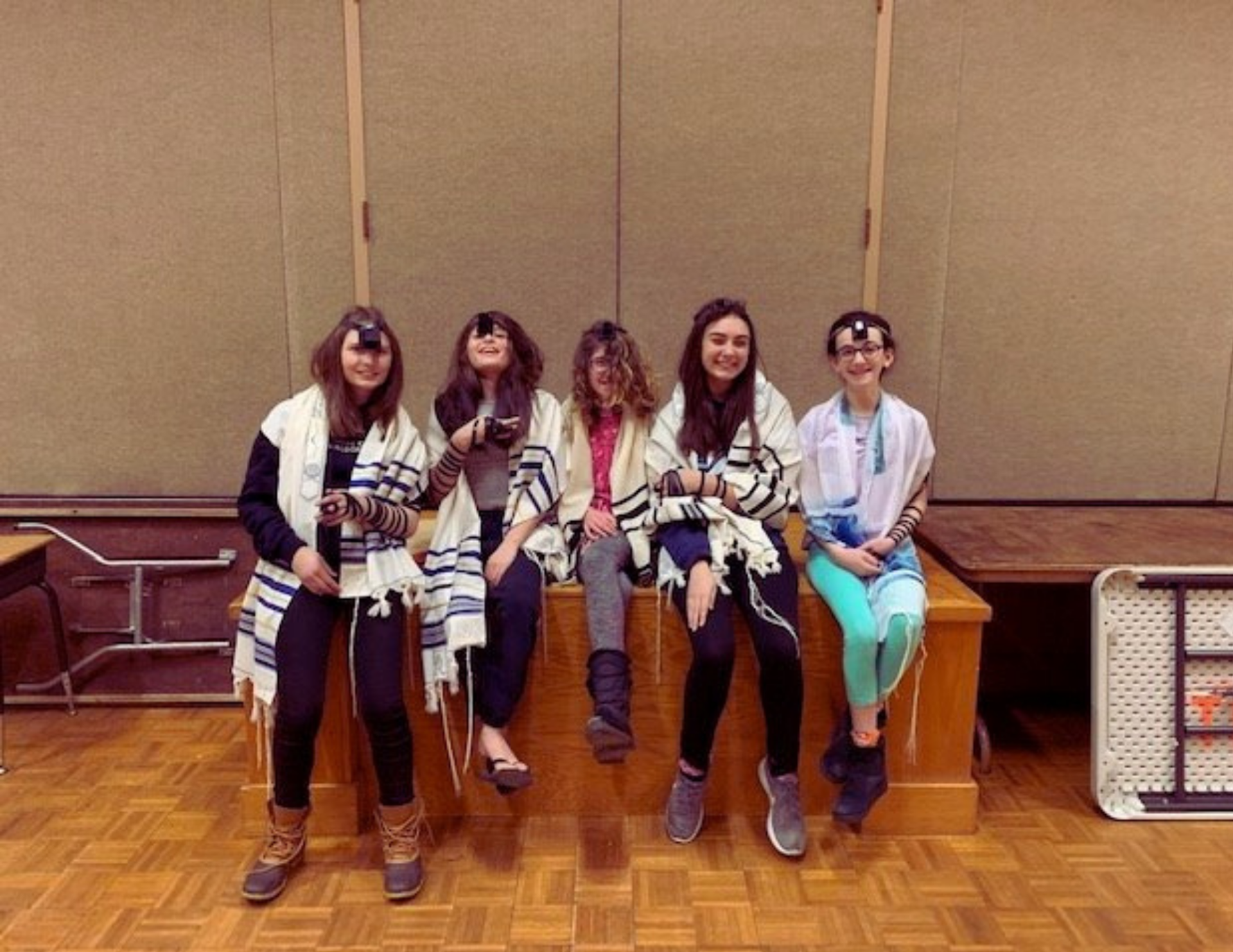 a photo of five bat mitzvah students, smiling at the camera, wearing tallitot and tefillin