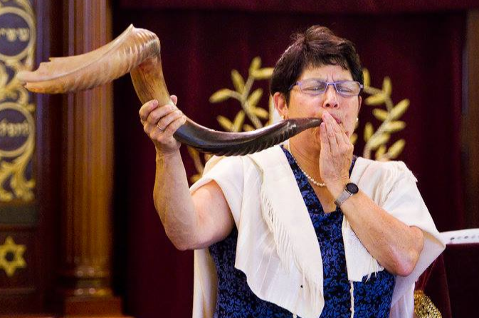 A photo of Rabbi Braun, a white woman with short hair, wearing a white tallis and blowing a large shofar