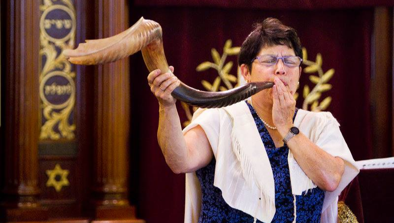 A photo of Rabbi Braun, a white woman with short hair, wearing a white tallis and blowing a large shofar