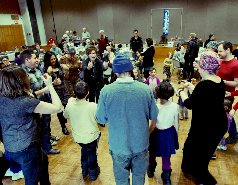 Many people in a circle in a synagogue's social hall, smiling and clapping