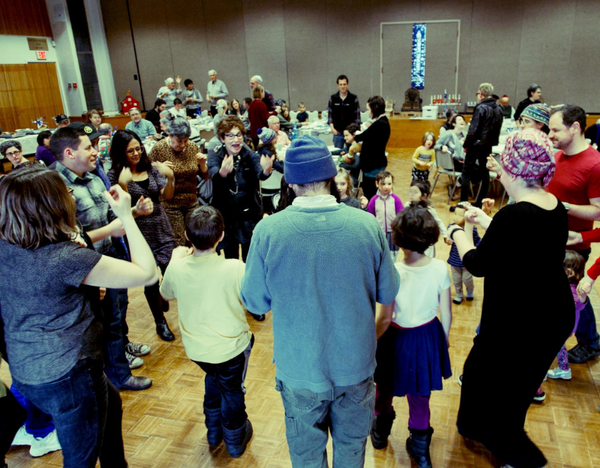 Many people are in a circle in a synagogue's social hall, smiling and clapping
