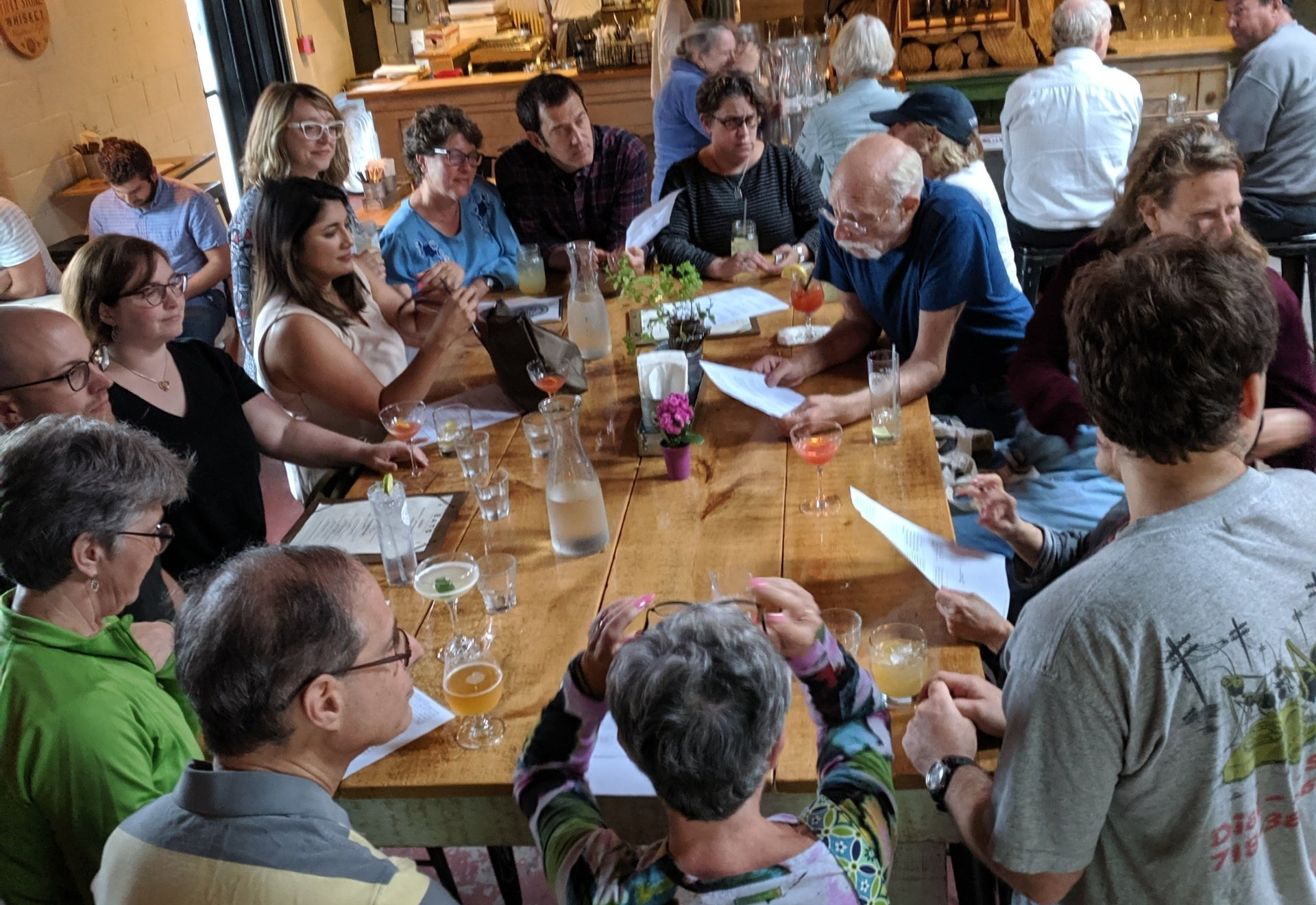 a photo of a group of adults (white, mostly middle aged) standing around a table at a bar, studying and talking to one another