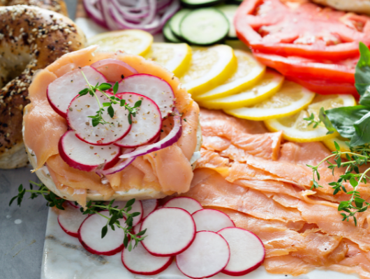 A photo of a bagel and lox spread with red onions, cucumbers, tomatoes, lemons, and radishes in the background