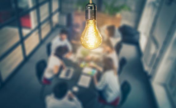 A stock photo of a bare lightbulb in the foreground, with people around an conference table behind it (out of focus)