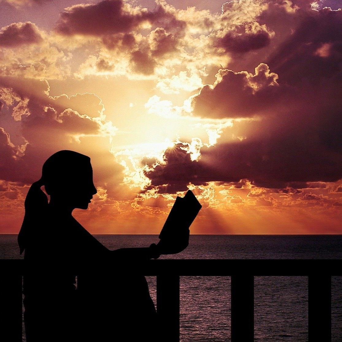 A photo of a person with a ponytail reading a book in silhouette against a sunset with lots of clouds above the ocean