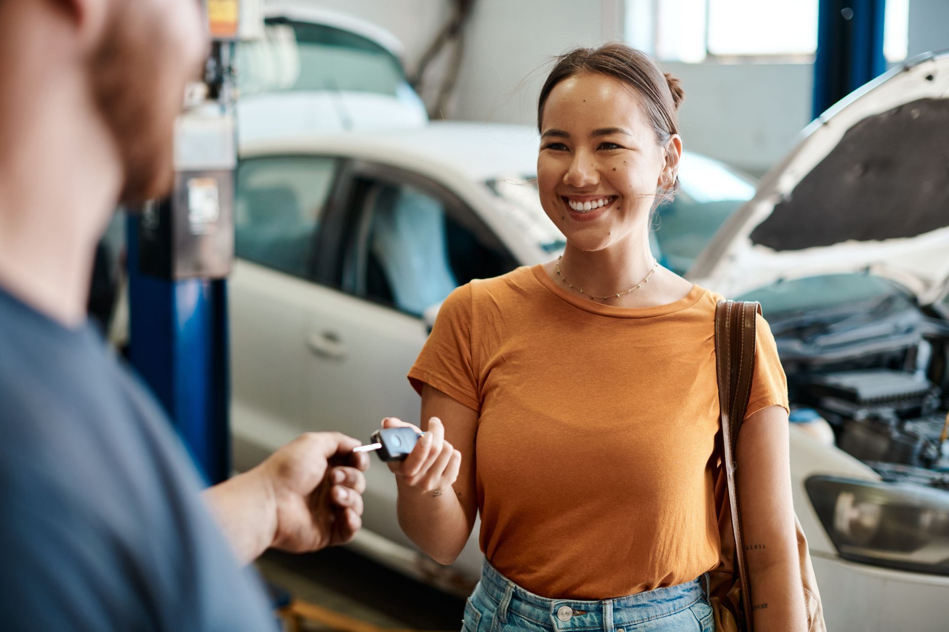 Customer hands over car keys at repair shop with white car being serviced in background.