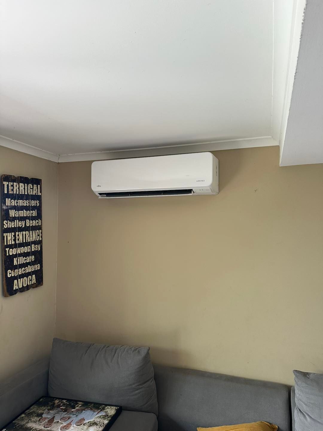 White Air Conditioner Mounted on a Tan Wall Above a Gray Sofa — DJL Refrigeration & Air Conditioning In Bowen, QLD