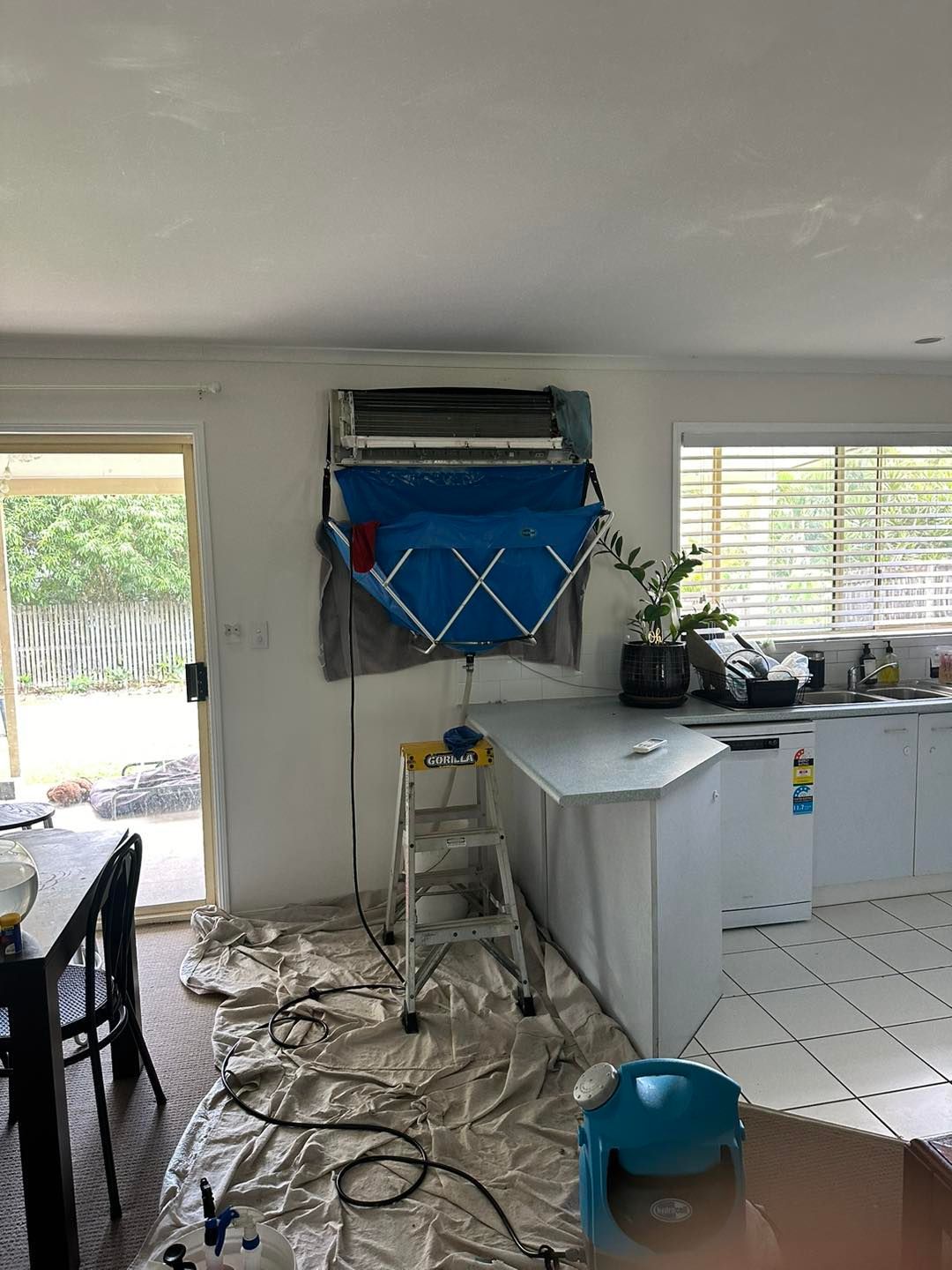 A split-system air conditioner with a blue cleaning bag attached, mounted above a kitchen counter, on a step ladder.