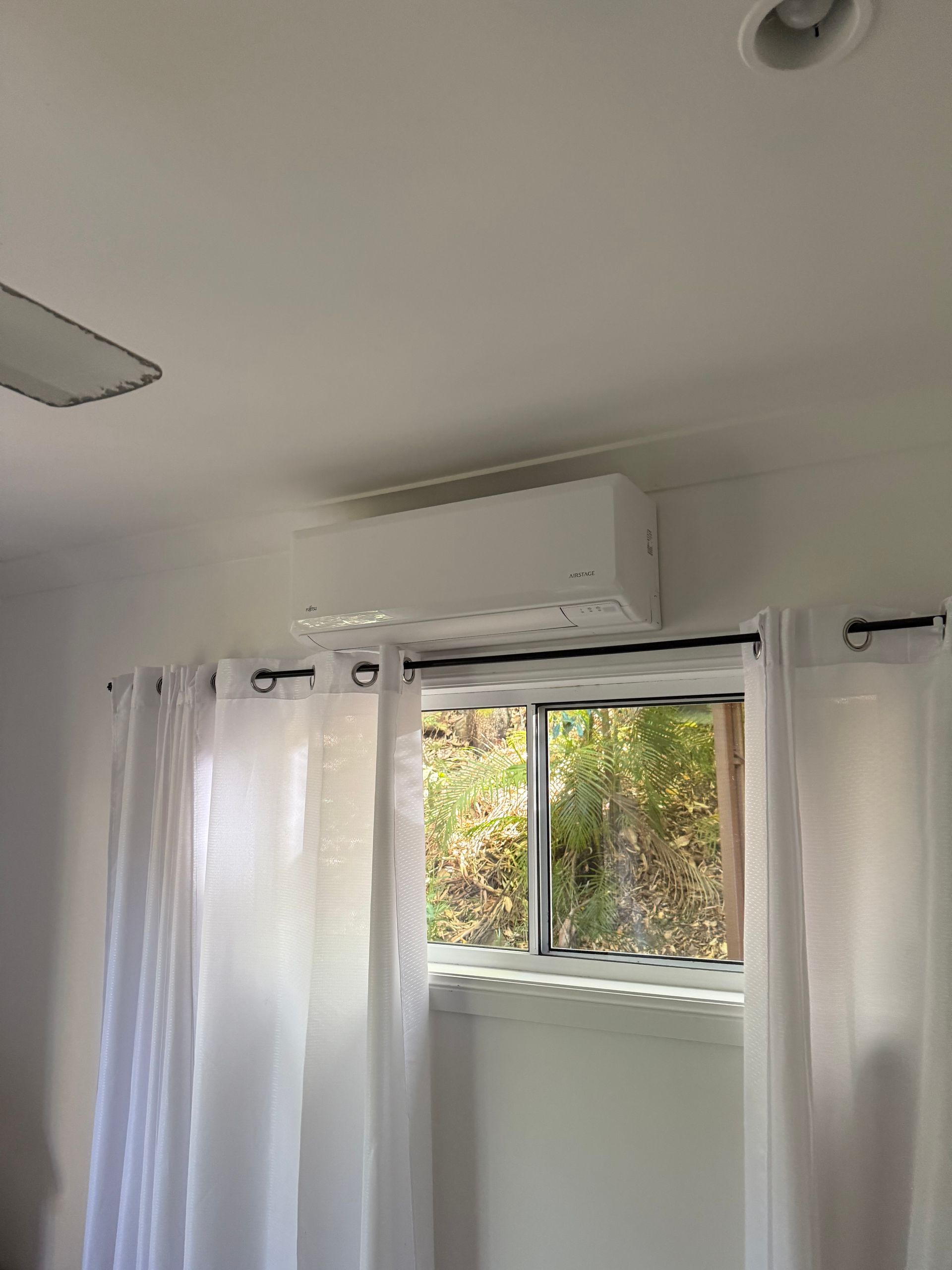 A white split-system air conditioner mounted on a wall above a window covered by sheer white curtains.