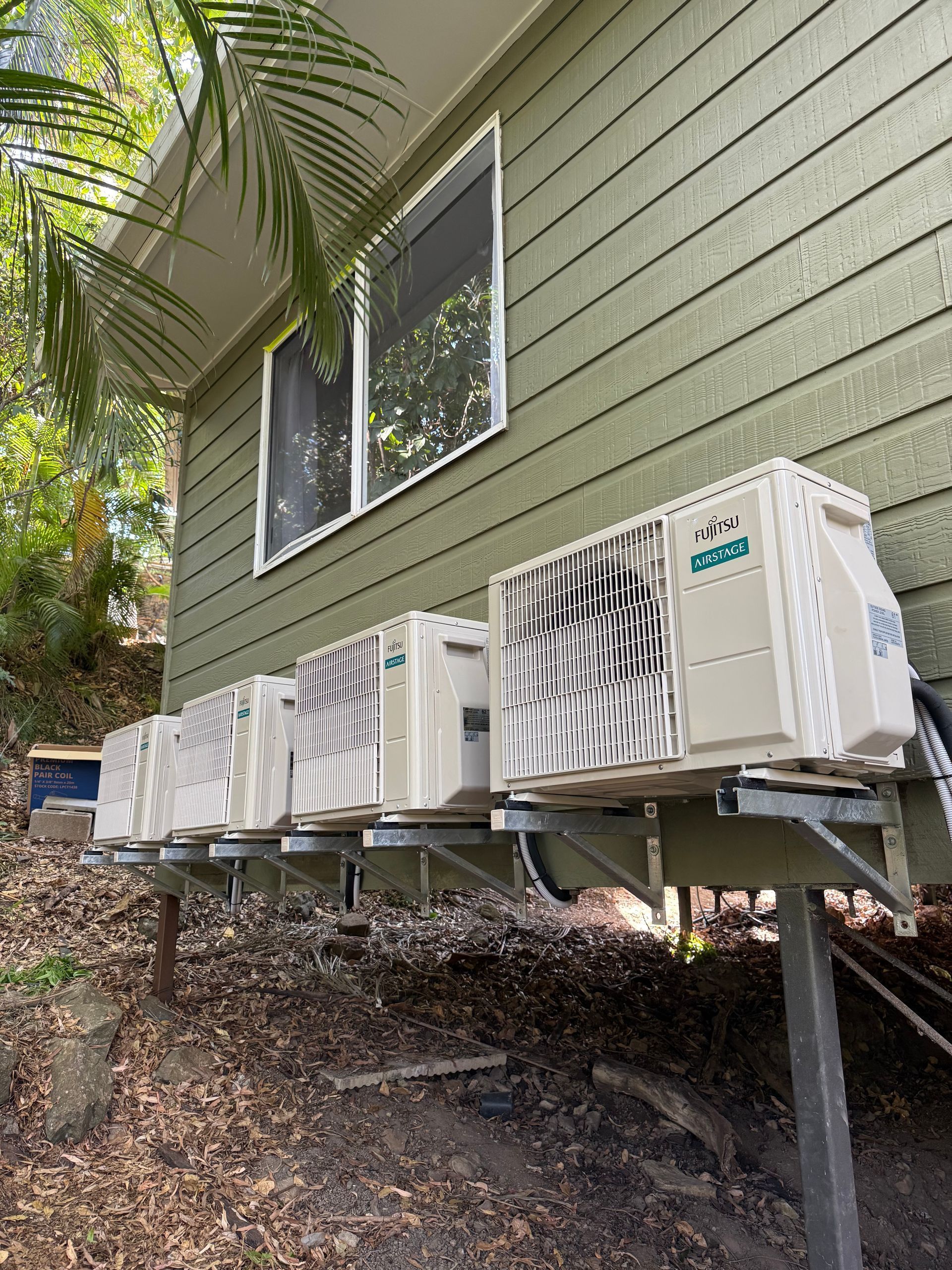 Four white air conditioning units mounted on metal brackets along the side of a green wooden house near a wooded area.