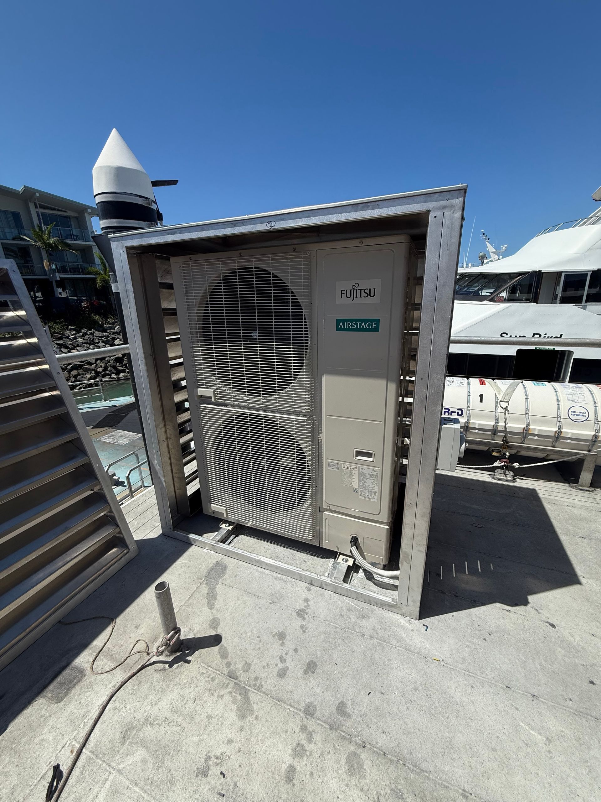 A beige air conditioning unit housed inside a protective metal enclosure on a concrete surface near a marina.
