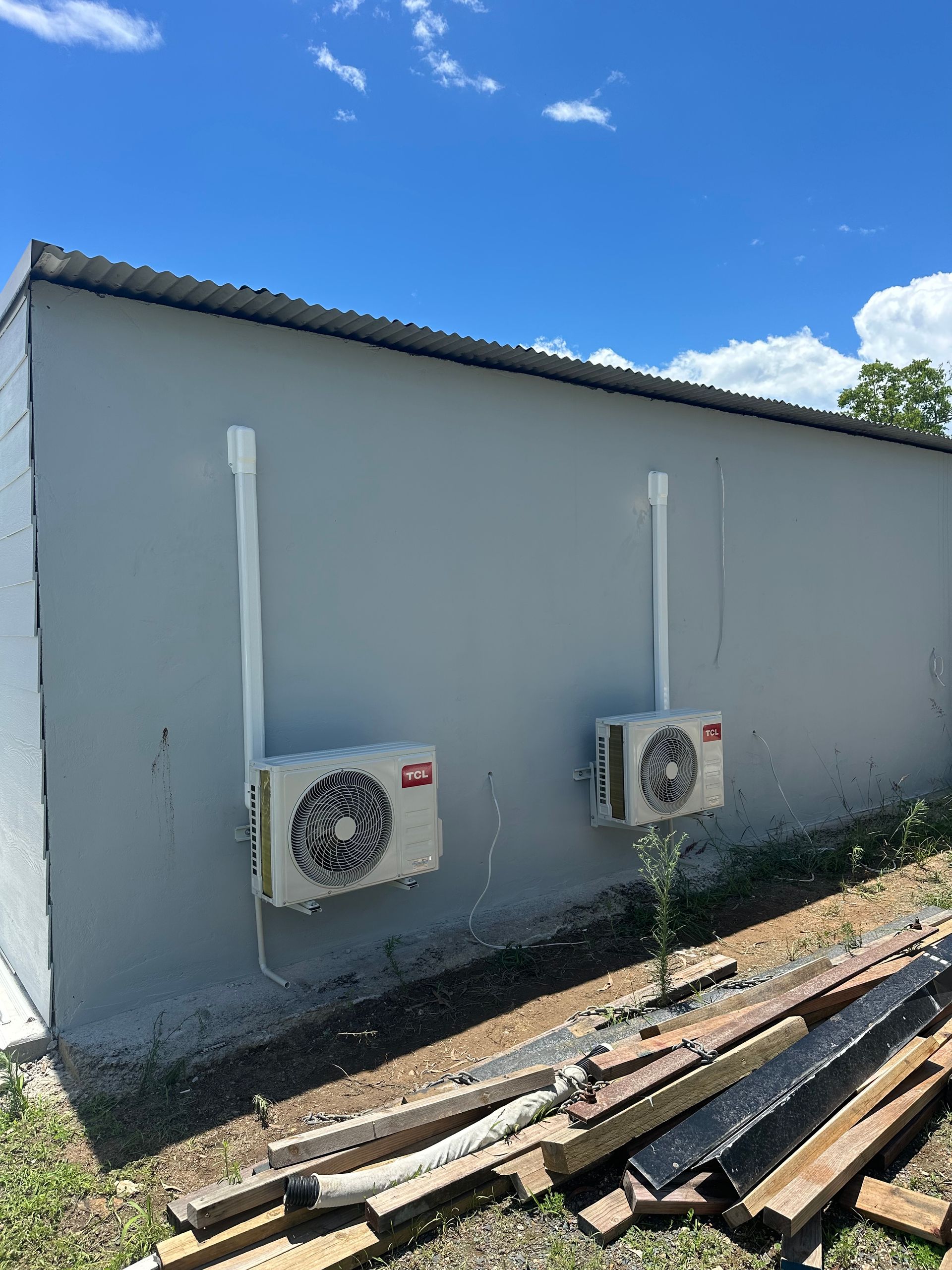 Gray building exterior with two AC units, white conduit pipes, and wood debris on the ground — DJL Refrigeration & Air Conditioning In Cannonvale, QLD