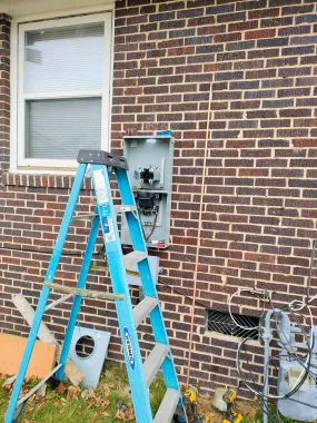 A ladder leans against a brick wall next to an electrical panel. A window is above the panel.