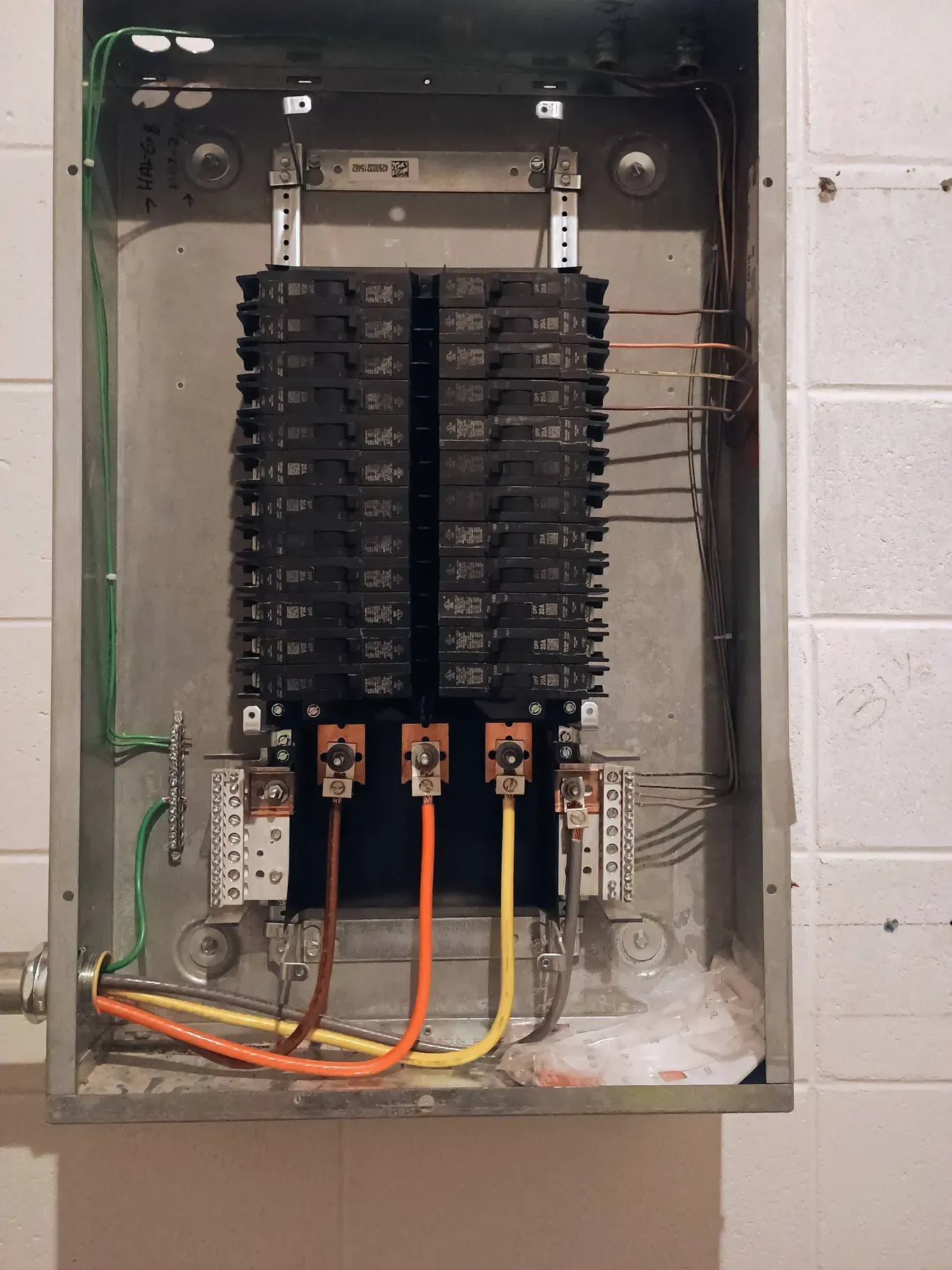 Electrical panel with rows of circuit breakers and colored wires. Mounted on a white tiled wall.