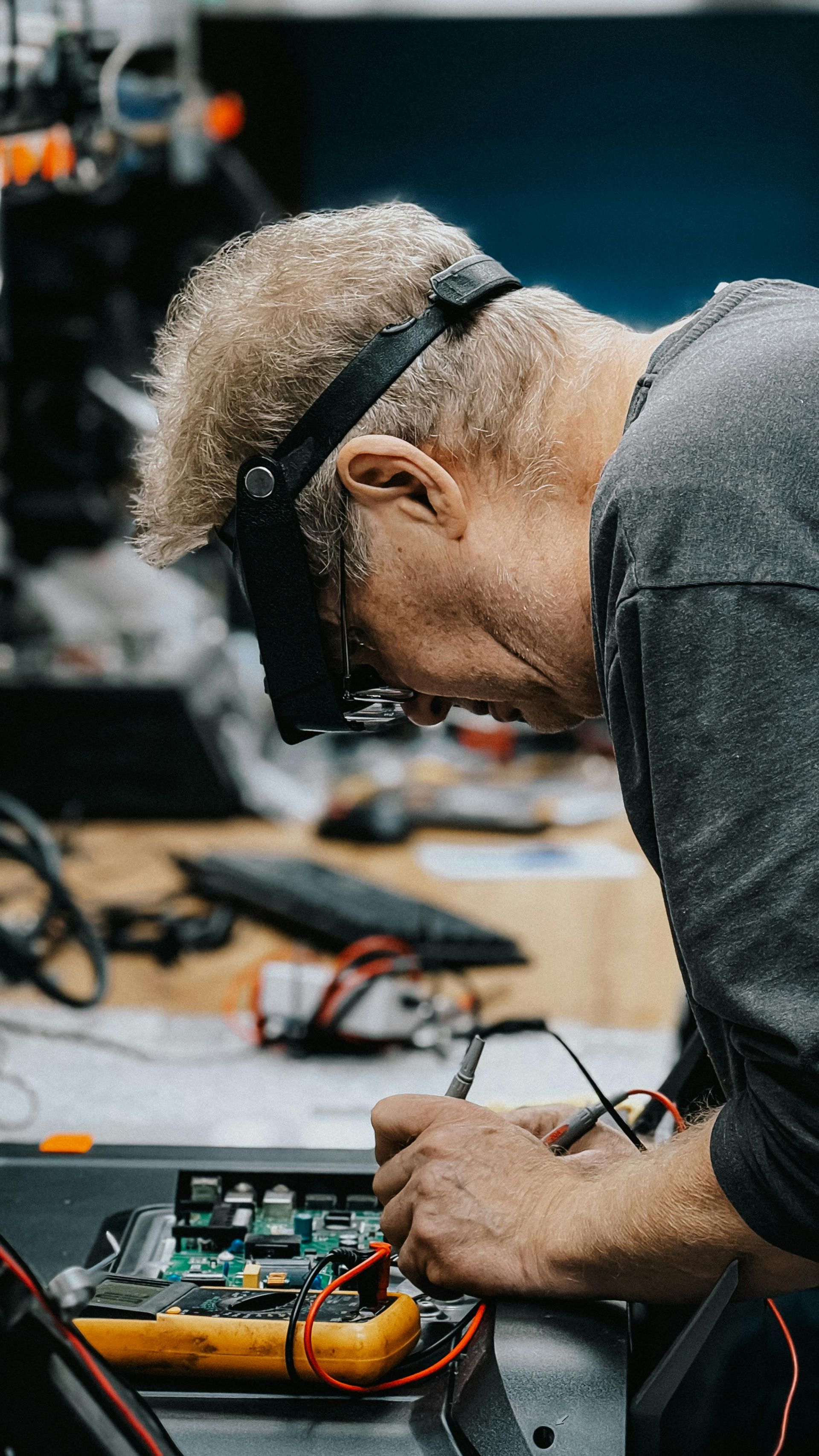 Person with magnifying visor inspects circuit board with multimeter. Indoors, focused work.