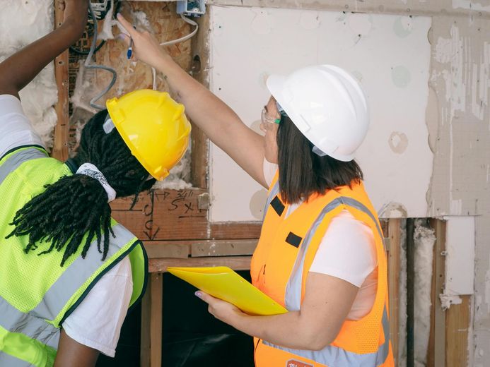 Two construction workers inspect electrical wiring inside a wall, one pointing with a yellow pad, both wearing vests and hard hats.