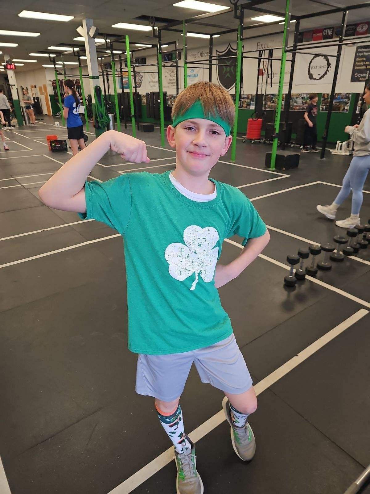 A young boy wearing a green shirt with a shamrock on it is flexing his muscles in a gym.