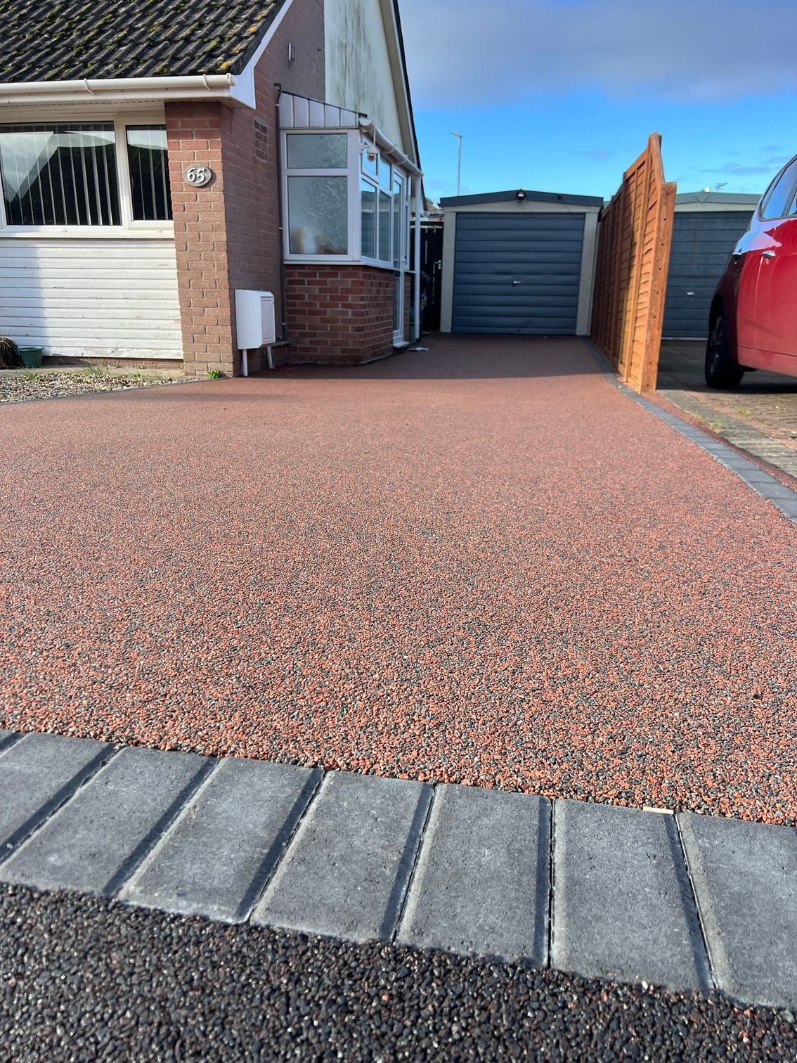 Red resin driveway with a grey block paving border, leading up to a black garage next to red brick.