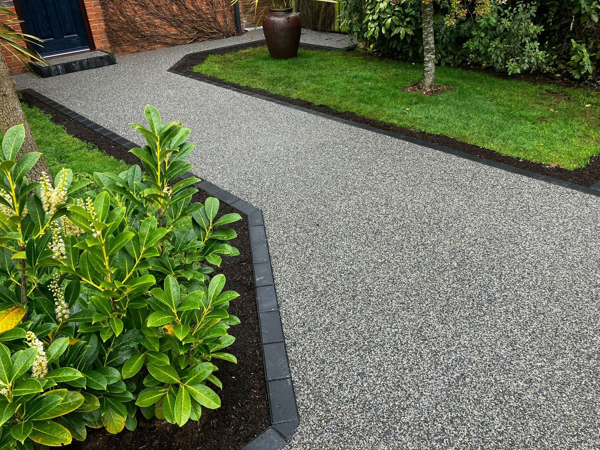 Grey resin driveway surrounded by dark block paving with green foliage.