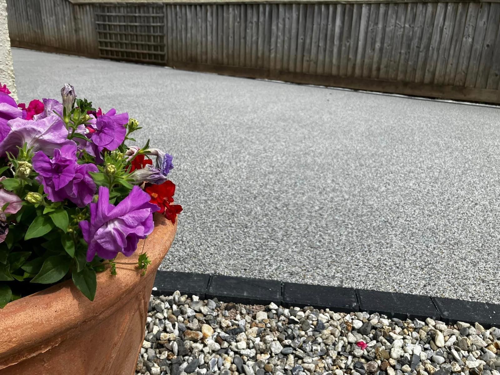 A pot of purple flowers is sitting on a gravel driveway.