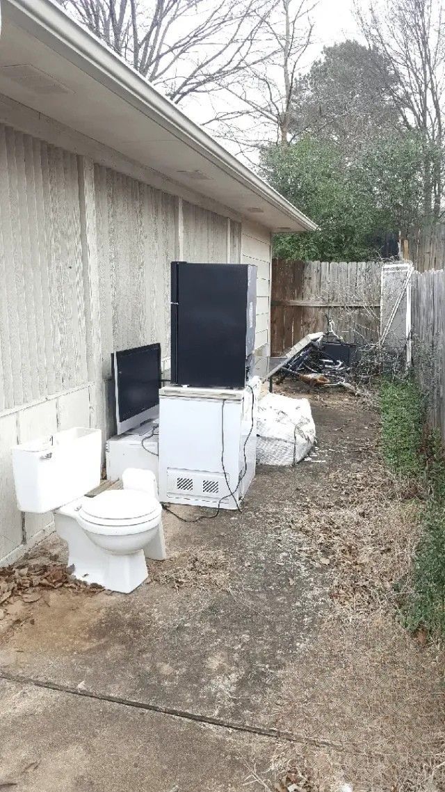 A toilet , refrigerator , television , and television are sitting outside of a house.