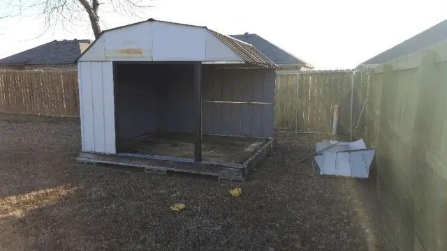 A small white shed is sitting in the middle of a dirt yard next to a fence.
