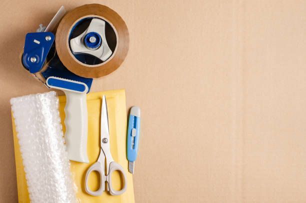 Packing supplies on cardboard: tape dispenser, bubble wrap, envelope, scissors, and a box cutter.