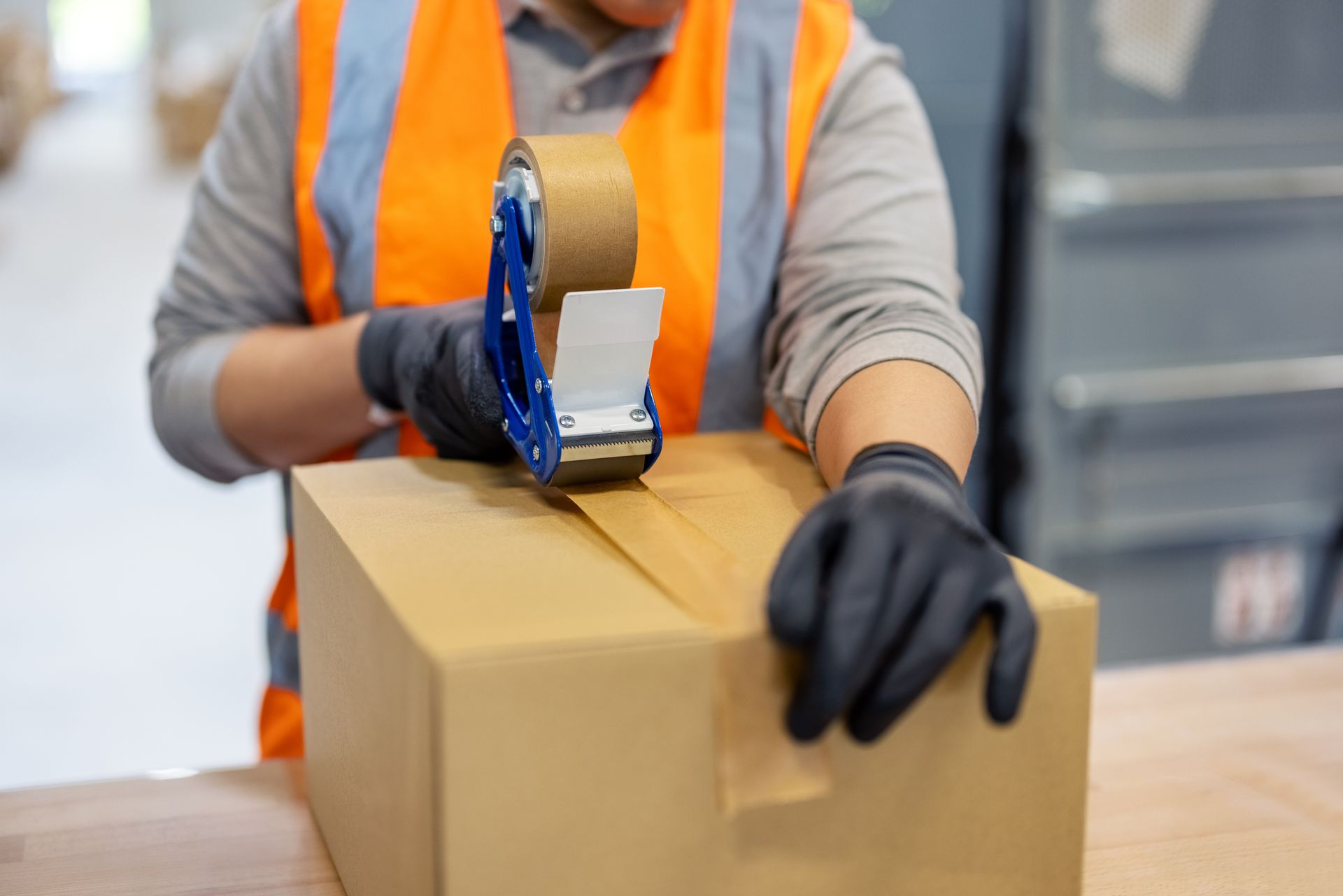Worker in safety vest seals a cardboard box with tape in a warehouse.