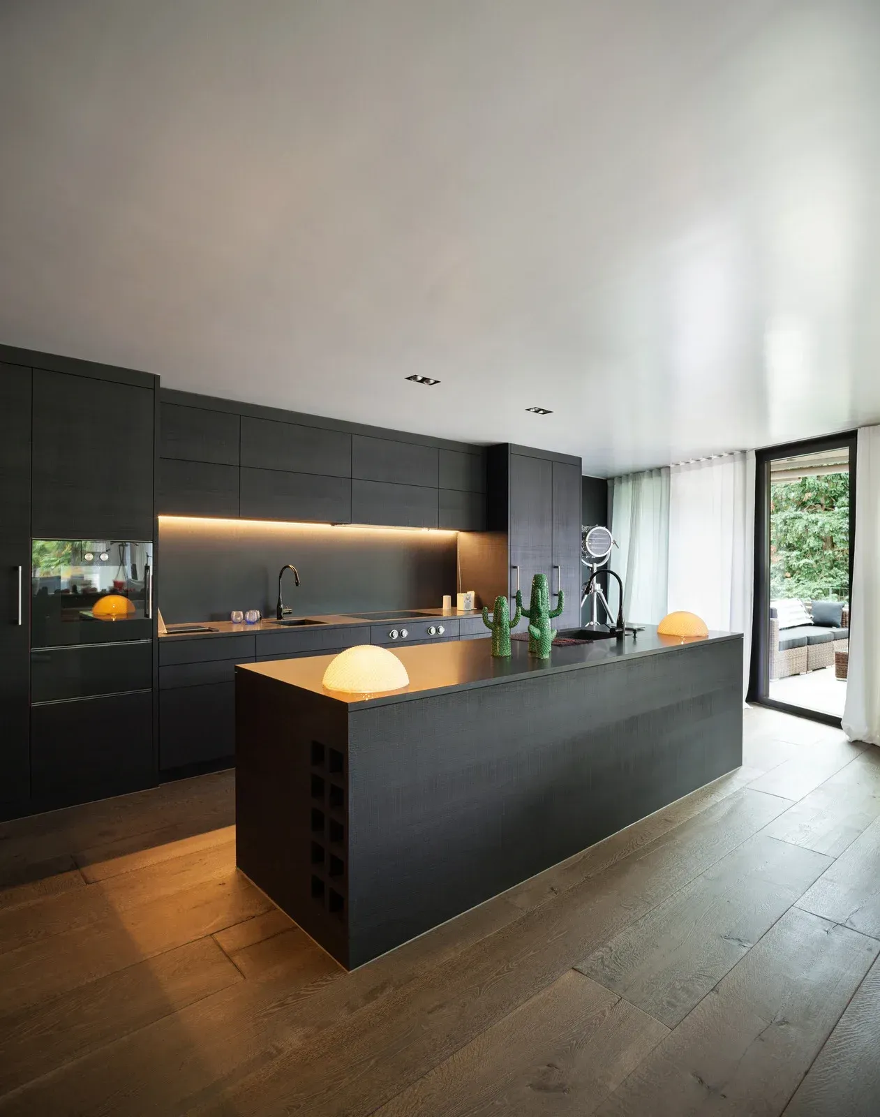Modern kitchen with black cabinetry, island, and wooden floors. Backlit counter, and outdoor view.