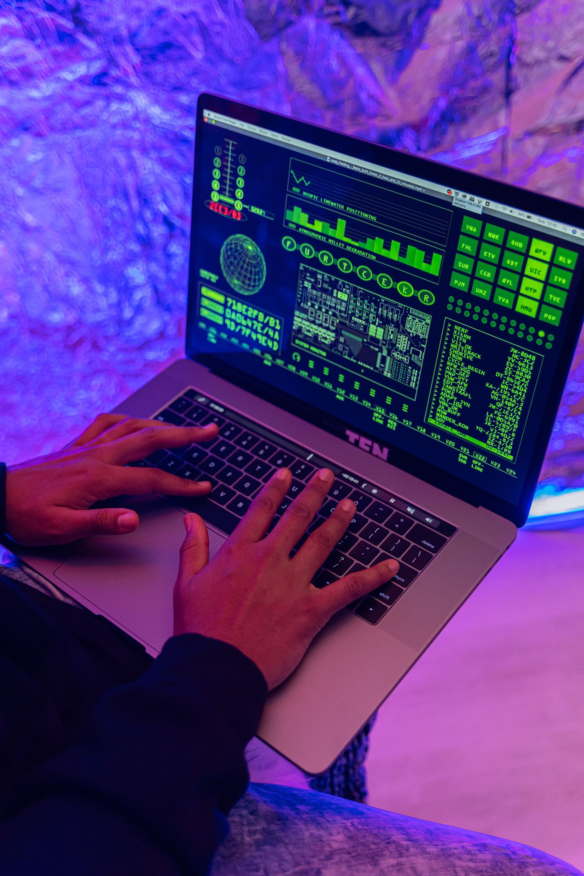 Hands typing on a laptop with green code on screen in purple-blue lighting