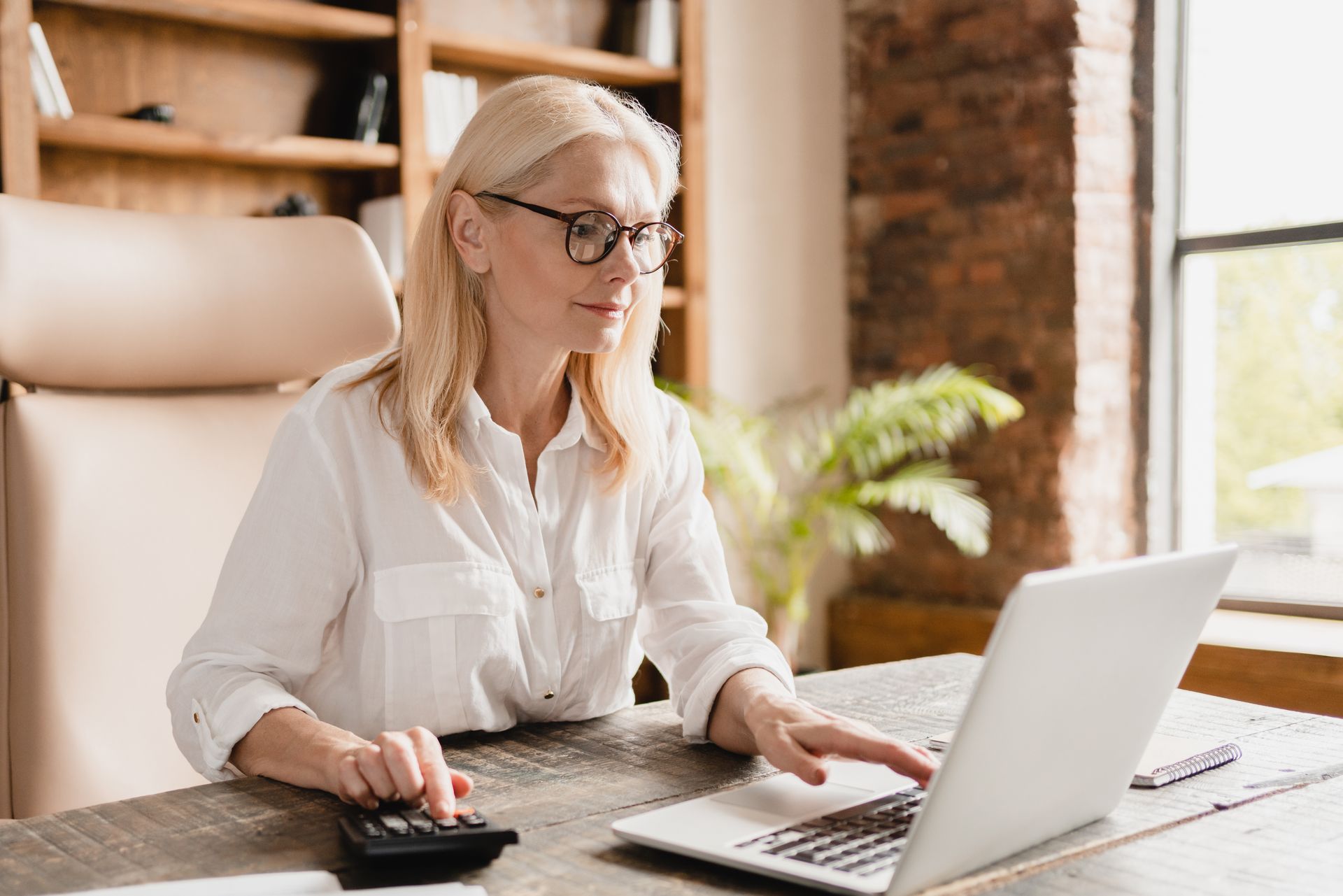 A woman is sitting at a desk using a laptop computer and a calculator.