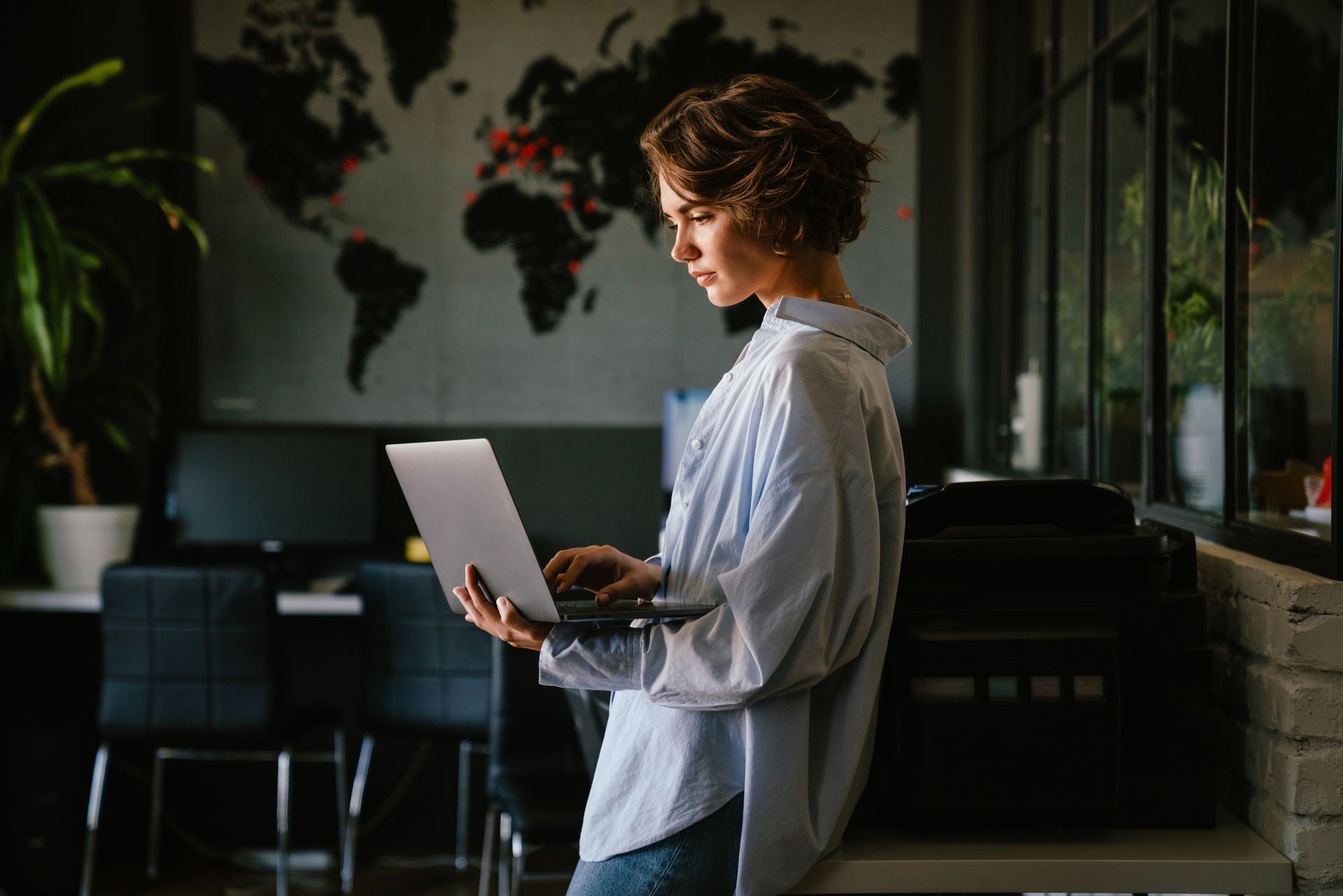 A person in a light blue shirt stands in an office, looking down at a laptop held in their hands.