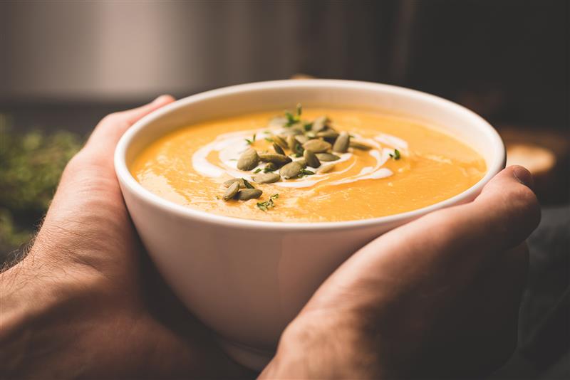 hands holding a bowl of orange-colored soup topped with herbs