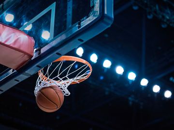 A picture of a basketball coming through the net with bright lights of the arena in the background.