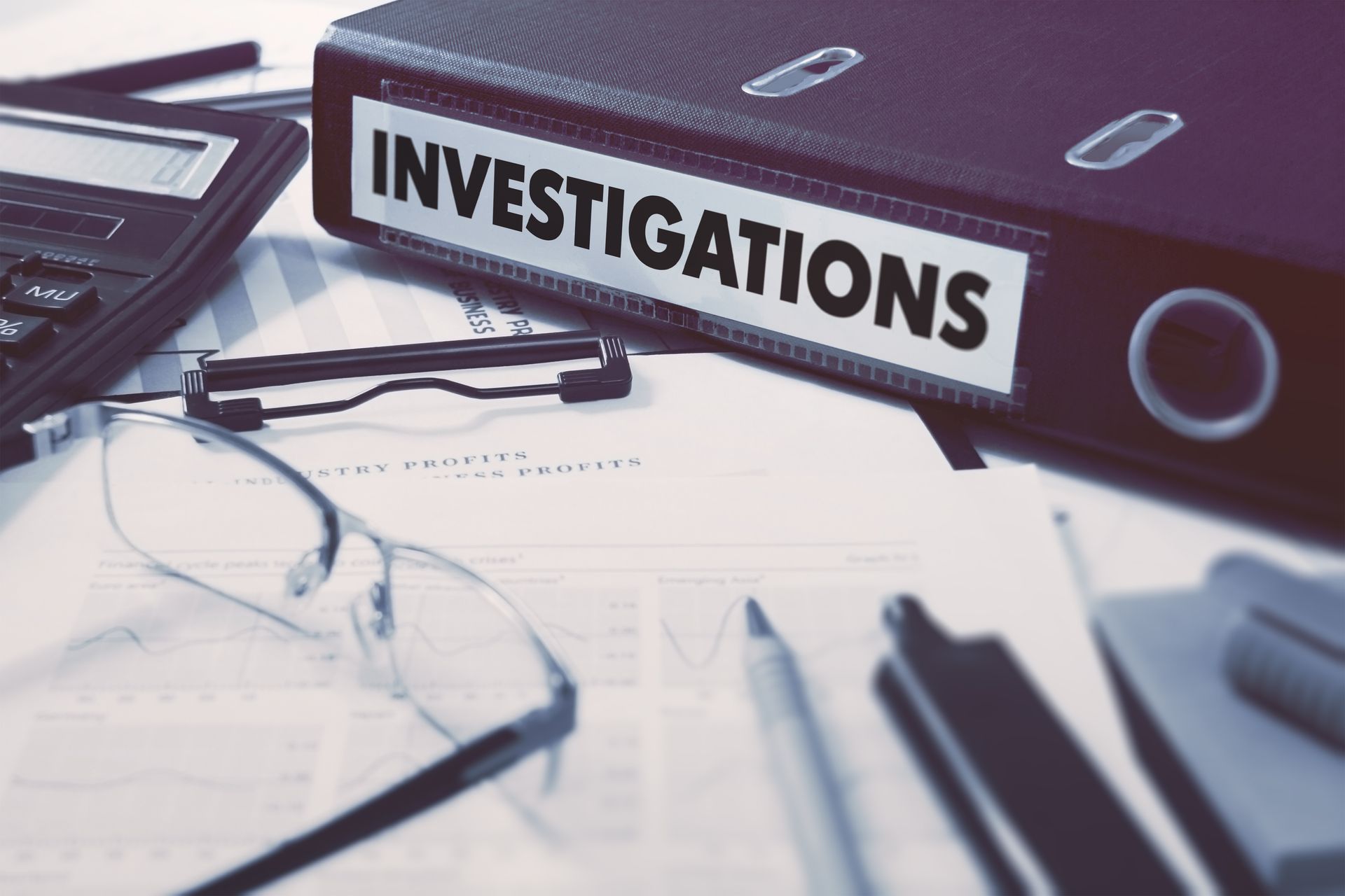 Binder labeled ‘Investigations’ on a desk with documents, glasses, and a calculator.